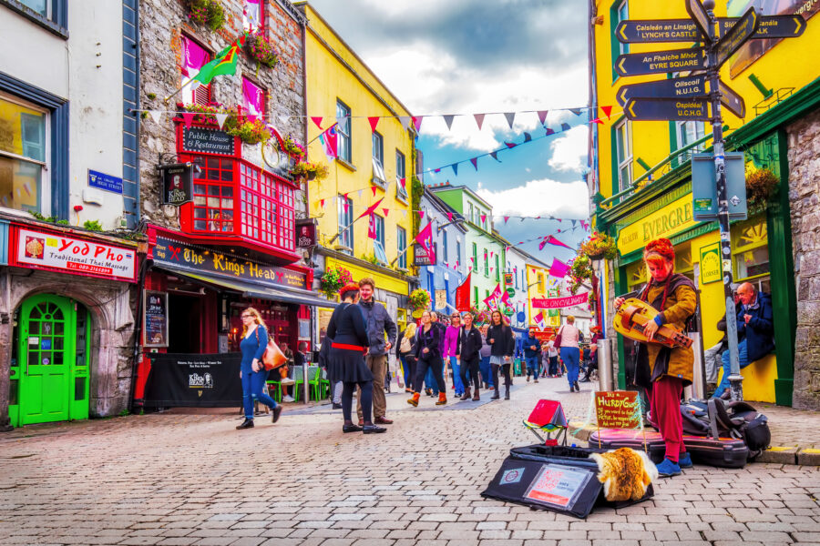 Galway Latin Quarter colorful cobbled street with busking musician, shops, and market crowds.