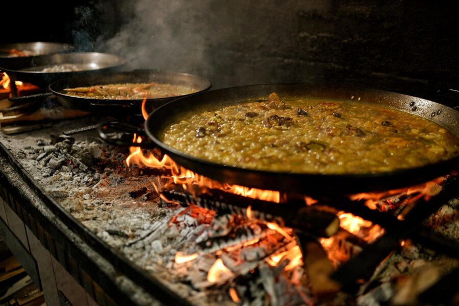 Rustic kitchen with open fire cooking, large pans, paella, traditional communal meal.