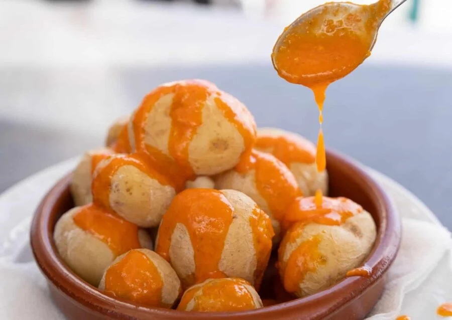 Boiled baby potatoes in a ceramic bowl with vibrant orange sauce being drizzled.