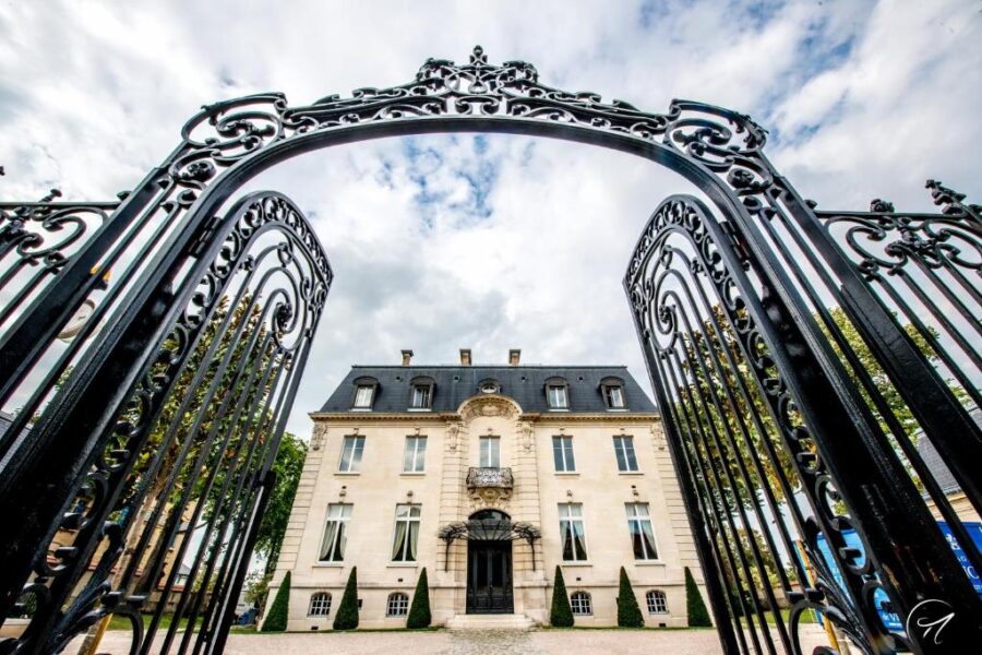 Ornate wrought-iron gates framing stately stone mansion entrance and gravel driveway.