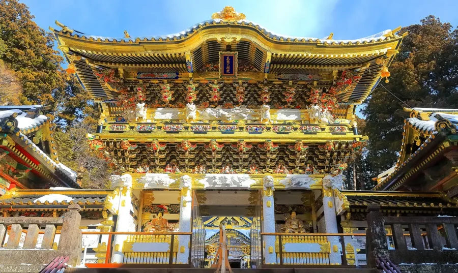 Ornate Nikko Toshogu Shrine gate in winter with gold details, snow-covered roof, and forest backdrop.