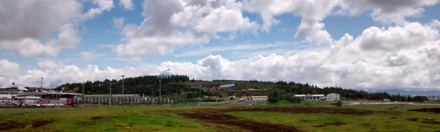 Scenic view from Öskjuhlíd Hill showing a grassy field, town structures, trees, and a vibrant, cloudy sky in the background.