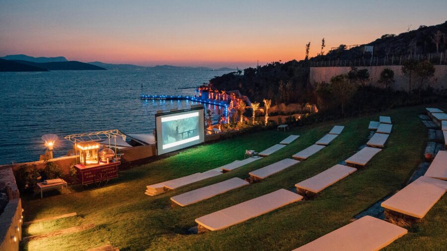 Outdoor waterfront cinema at sunset with loungers, screen, and snack cart.