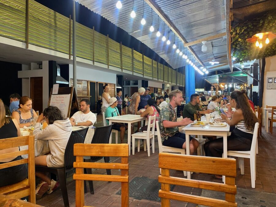 Outdoor restaurant with warm lighting, diverse diners enjoying a lively, cozy ambiance.