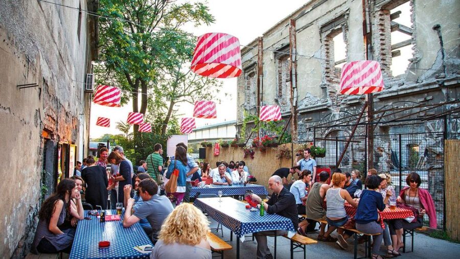 Outdoor dining scene with rustic tables, lively diners, urban decay, and greenery in Savamala District.
