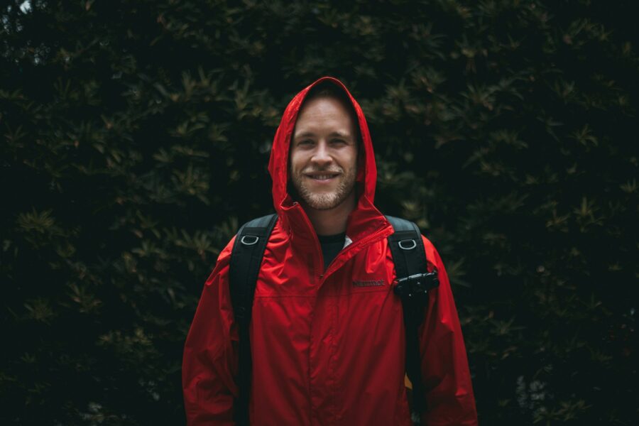Young man in red rain jacket smiling, ready for adventure against lush green foliage.