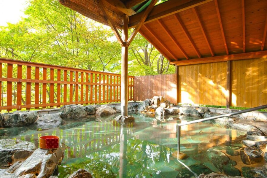 Outdoor Japanese onsen hot spring bath with rocks, forest views and traditional wooden shelter.