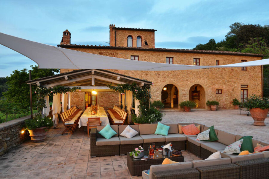 Rustic outdoor lounge and dining area with stone building, colorful pillows, and lush greenery.