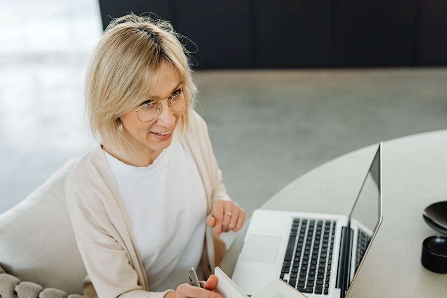 Blonde woman with glasses using laptop and notebook in modern remote work workspace.