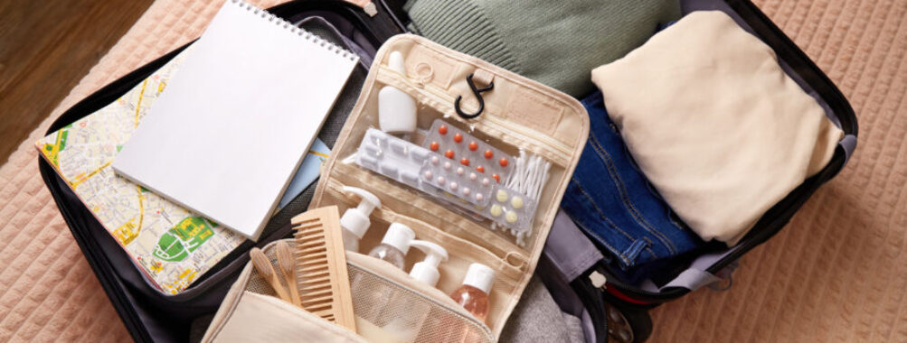 Open suitcase with neatly packed clothes, a green sweater, beige shirt, jeans, a toiletry bag, notebook, and a map. It conveys readiness for travel.