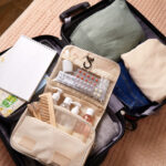 Open suitcase with neatly packed clothes, a green sweater, beige shirt, jeans, a toiletry bag, notebook, and a map. It conveys readiness for travel.