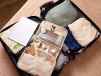 Open suitcase with neatly packed clothes, a green sweater, beige shirt, jeans, a toiletry bag, notebook, and a map. It conveys readiness for travel.