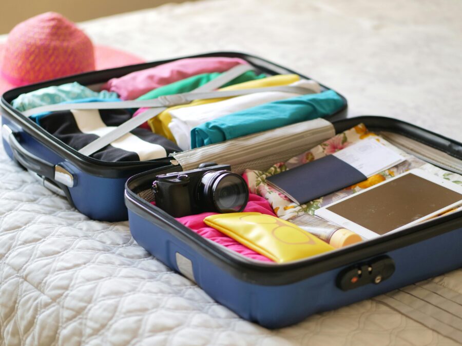 Open suitcase on bed with colorful clothes, travel essentials, and sunhat, suggesting vacation preparation.
