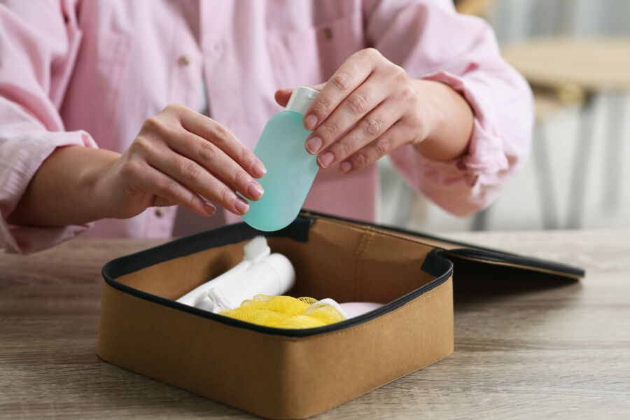 Woman organizing a cosmetic travel kit into a compact toiletry bag on a wooden table, with bath accessories visible.
