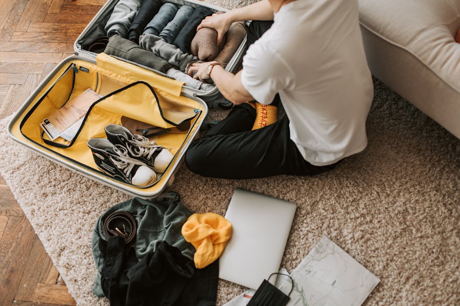 Person packing suitcase with clothes and laptop, preparing for travel on a cozy rug.