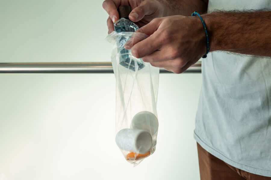 Person holds a clear bag containing a cracked egg shell and two whole eggs over a railing, suggesting caution. The background is softly lit.