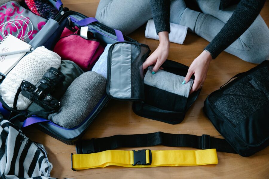 Person packing suitcase with clothes, camera, and notebook for a trip.