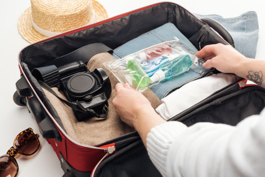 Person packs a red suitcase, placing a clear toiletry bag on top of folded clothes, next to a camera. A hat and sunglasses lie beside the suitcase.