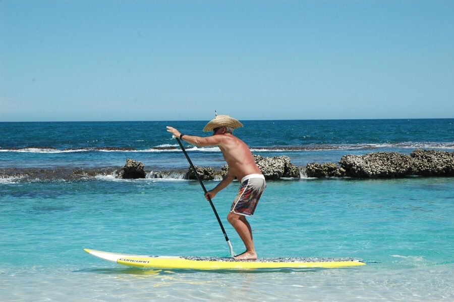 Man paddleboarding on bright yellow board in calm turquoise sea, wearing straw hat.