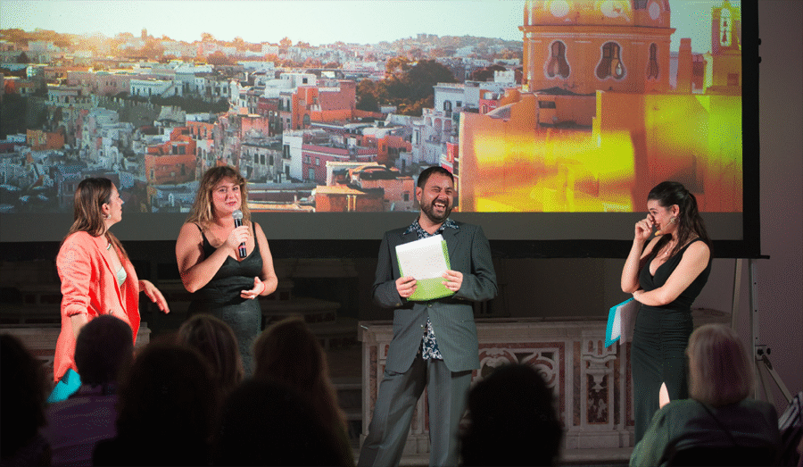 Procida Film Festival panel discussion with presenters in front of sunlit city projection.