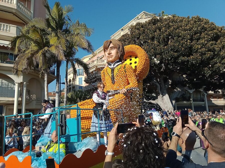 Orange figure float in festive parade with crowd, palm trees, and confetti.