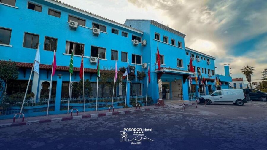 Bright blue Parador Hotel in Chefchaouen with flags, parking lot, and exterior mountain logo.