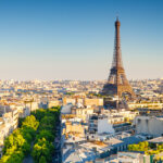 Aerial view of Paris during sunset, featuring the Eiffel Tower prominently. The skyline is bathed in warm light, with green trees lining the streets.