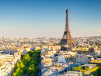 Aerial view of Paris during sunset, featuring the Eiffel Tower prominently. The skyline is bathed in warm light, with green trees lining the streets.
