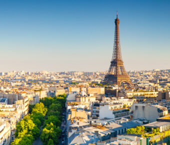 Aerial view of Paris during sunset, featuring the Eiffel Tower prominently. The skyline is bathed in warm light, with green trees lining the streets.