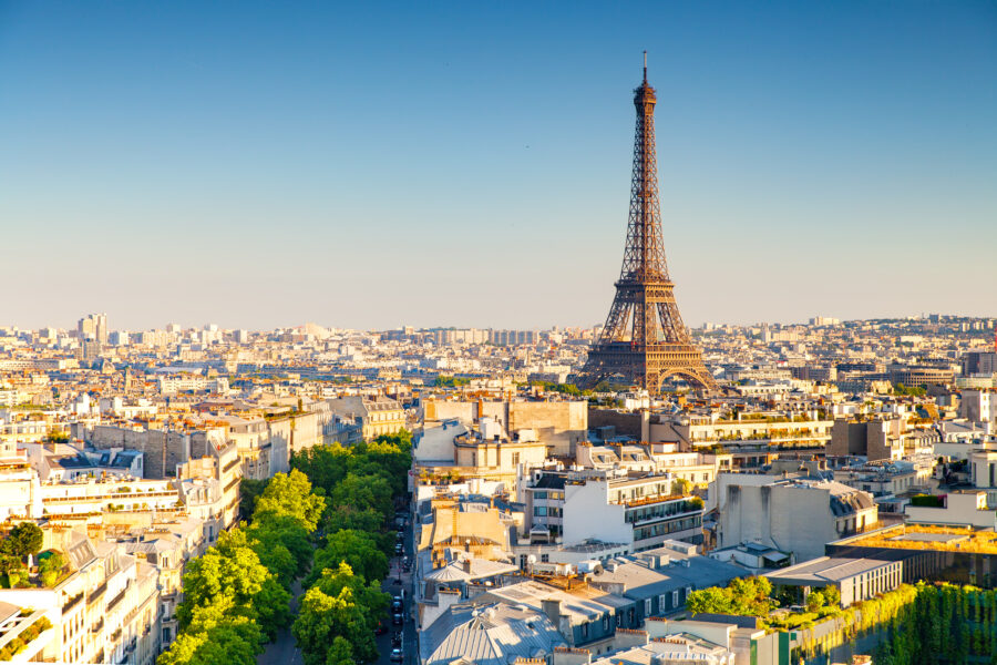 Aerial view of Paris during sunset, featuring the Eiffel Tower prominently. The skyline is bathed in warm light, with green trees lining the streets.