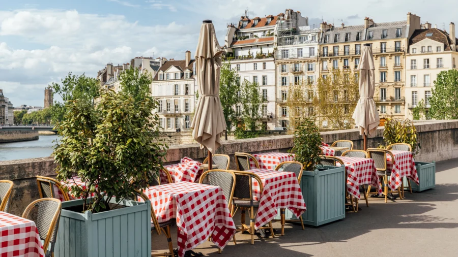 Charming Parisian waterfront bistro with checkered tables, wicker chairs, lush greenery, and Haussmannian buildings.