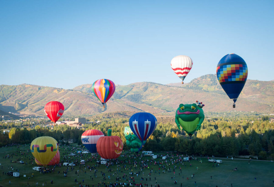 Colorful hot air balloons float over Park City during autumn, surrounded by vibrant fall foliage and clear blue skies.