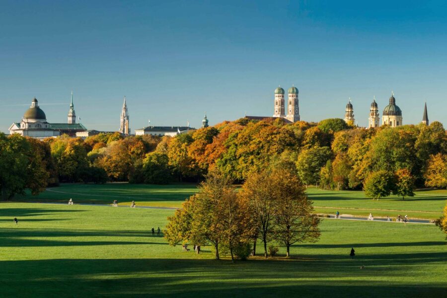 Autumn park lawn with people walking and historic towers skyline under clear blue sky.