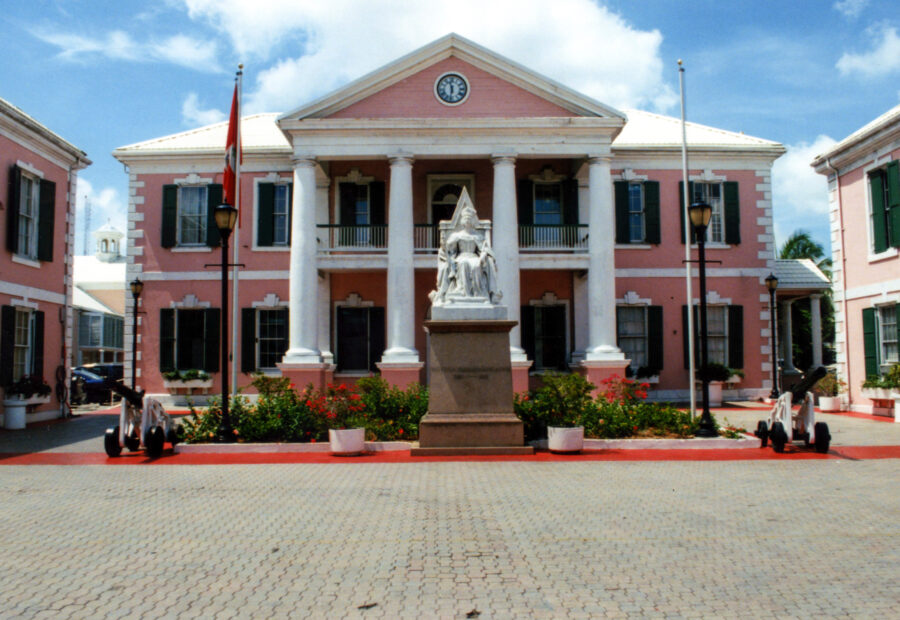 Parliament Square Nassau colonial building with white columns and a statue in front, surrounded by green plants and flags. Bright day, blue sky with clouds.