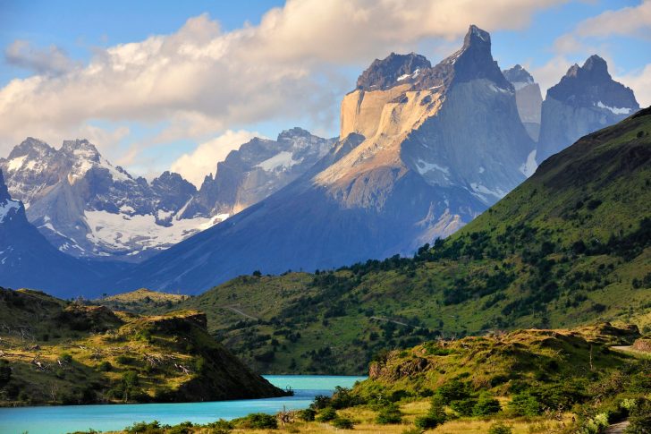 Patagonian mountain lake landscape with turquoise water, green hills, snow-capped peaks and dramatic sky.