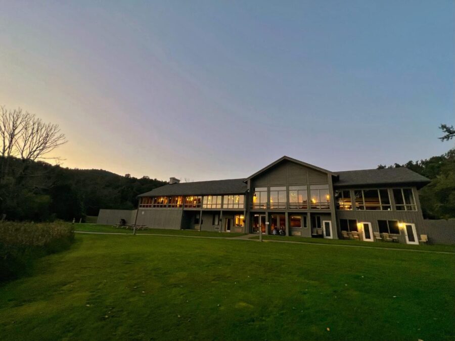 Large, two-story building with glass windows glows warmly at dusk in Peaks of Otter Virginia . It stands surrounded by a green lawn and trees, evoking a peaceful, inviting atmosphere.