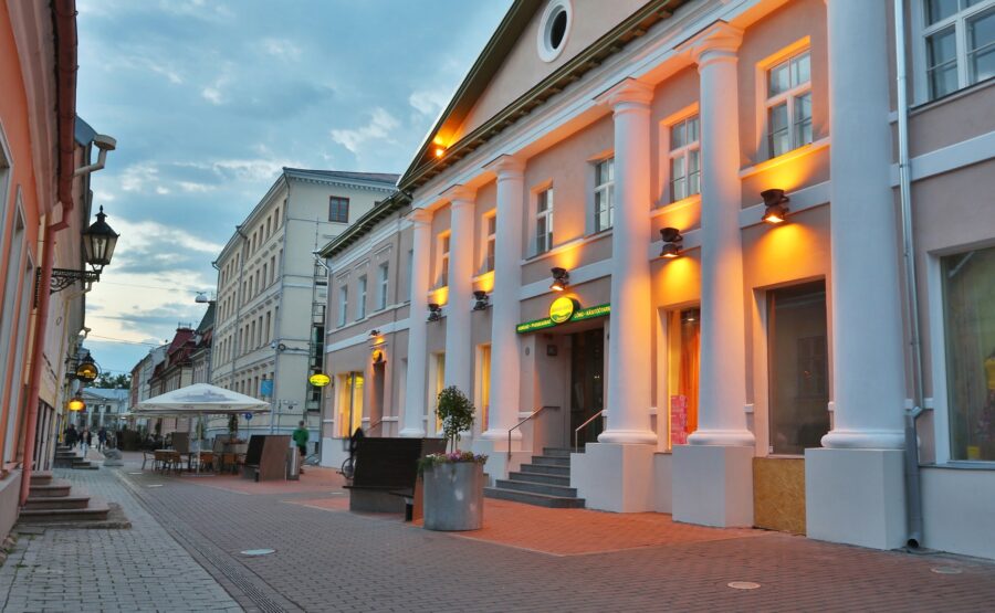 Evening pedestrian street in Tartu with classical columns, outdoor cafe seating, warm architectural lighting.