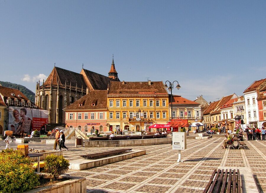 People stroll through Piața Sfatului in Brașov, featuring a vibrant city square and a prominent church.