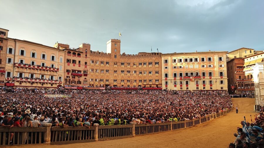 Crowded Palio arena in historic Italian piazza with sand track and medieval buildings.