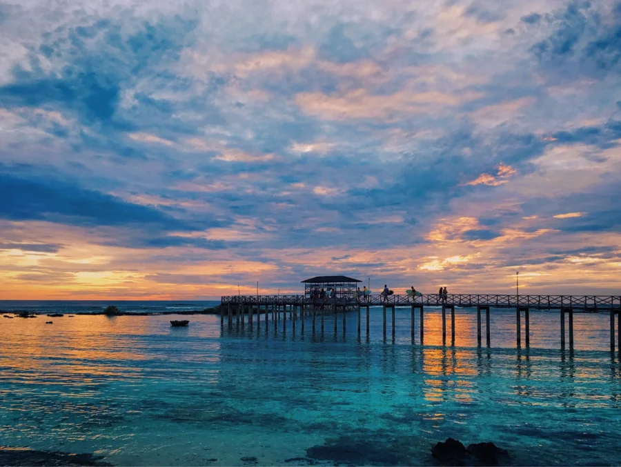 Wooden pier at sunset over turquoise seascape with silhouetted figures and dramatic clouds.