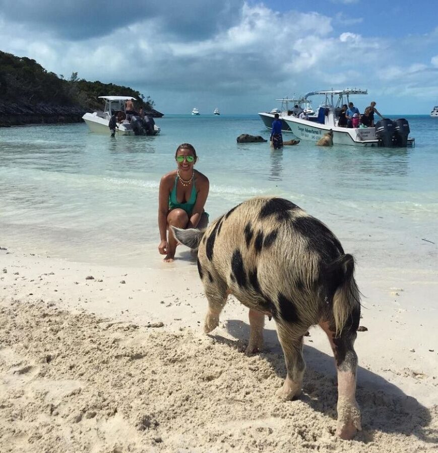 Woman kneels beside a pig on the beach during the Pig Beach Bahamas Pig Experience.