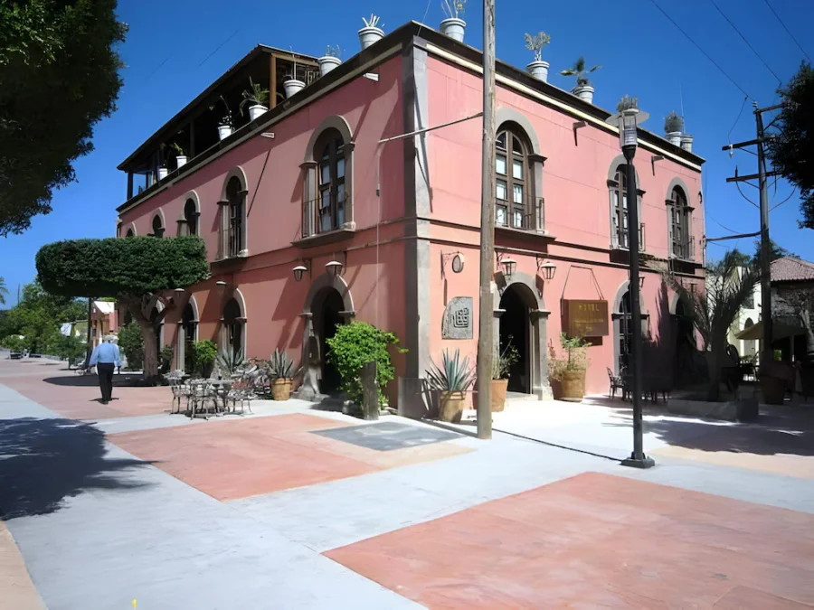 Pink colonial-style hotel on sunny Mexican street corner with terrace, arches, palms and plaza.