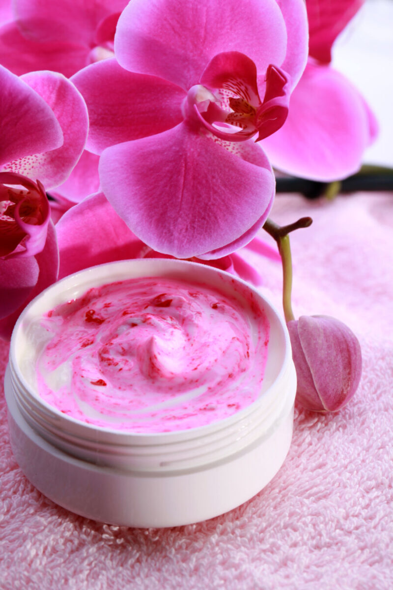 Pink orchid flowers beside an open jar of swirled pink cream on a soft, pink textured fabric. The scene is bright and serene.
