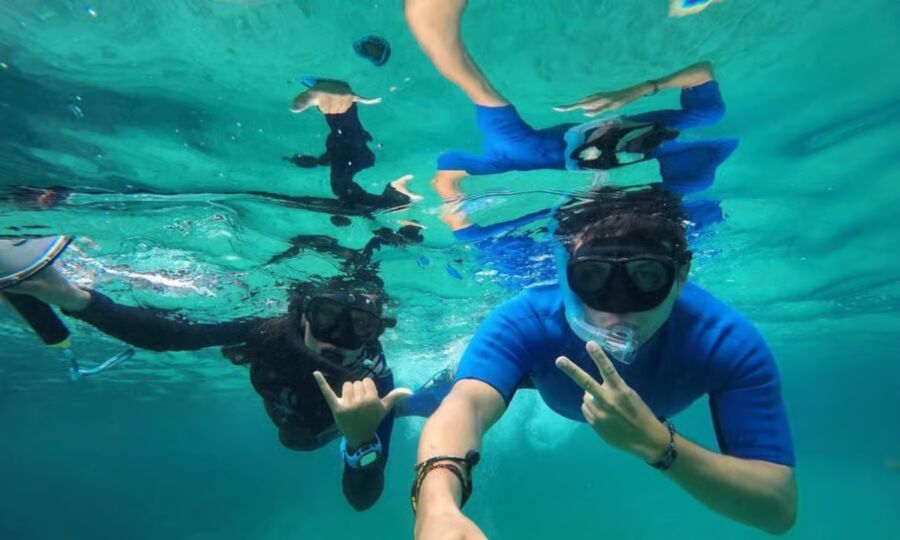 Two people snorkeling underwater, wearing masks and snorkels. They make playful hand gestures, surrounded by clear blue ocean water, creating a joyful vibe.