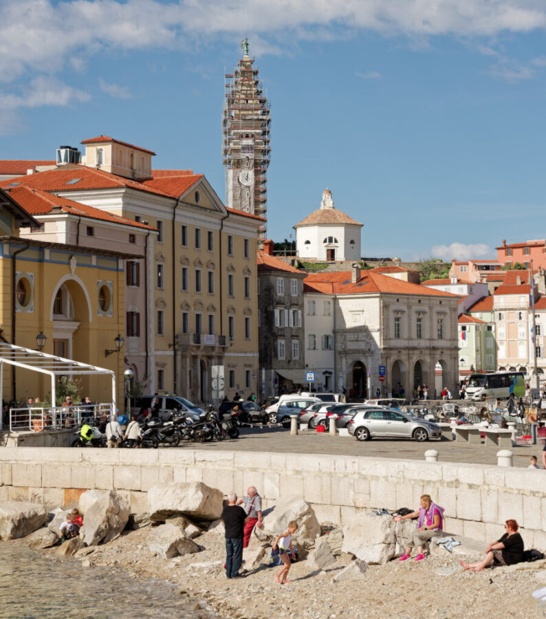 Coastal Piran, Slovenia Town scene with people relaxing on a rocky beach in front of historic buildings.