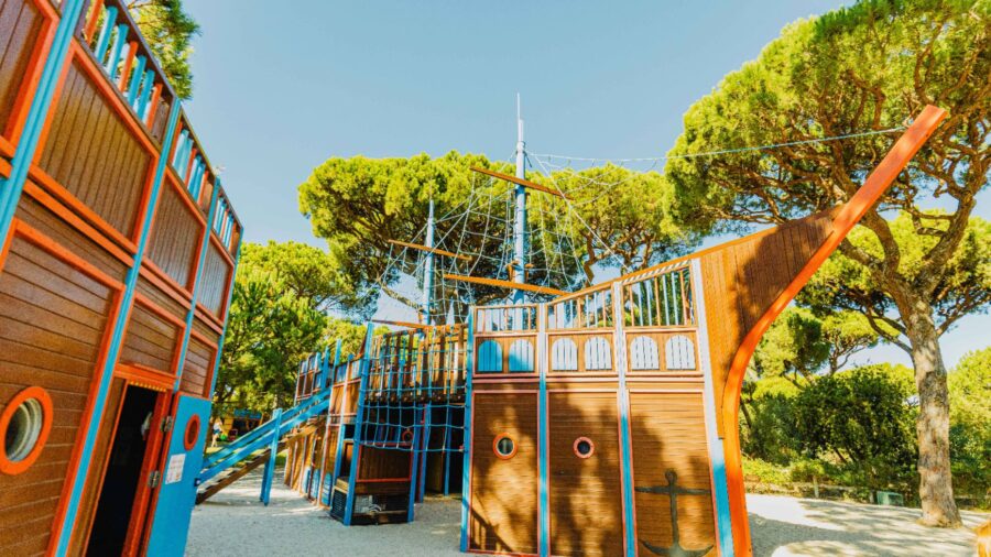 Pirate ship playground with wooden structure, colorful accents, climbing ropes, slides, and lush trees.