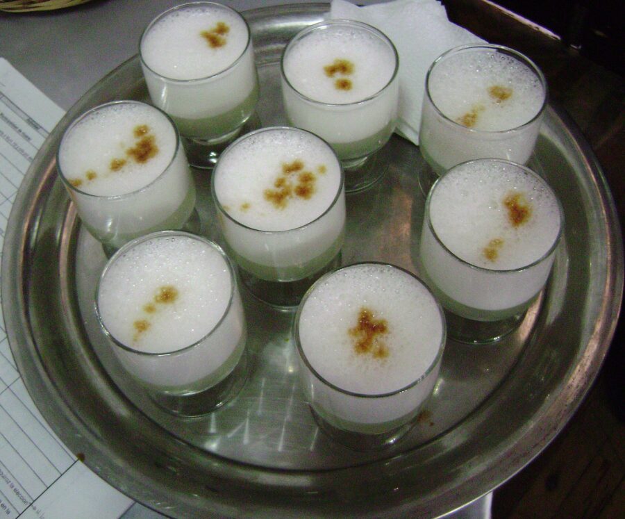 Six glasses of pisco sour, a creamy white cocktail, are arranged on a silver tray, ready for serving.
