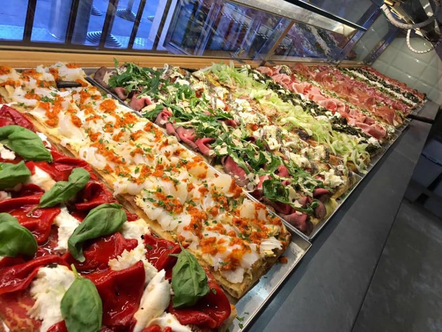 Gourmet pizza al taglio display case with rectangular pizza slices and assorted toppings.