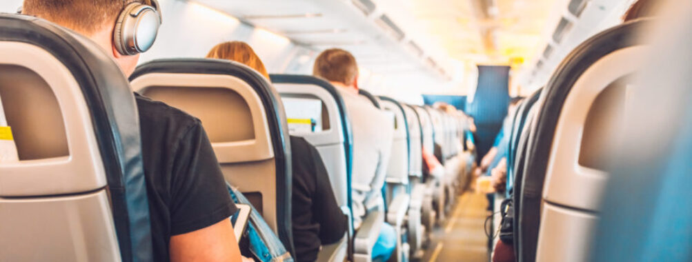 Economy class airplane cabin with passengers seated in armchair-style seats along a central aisle.