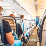 Economy class airplane cabin with passengers seated in armchair-style seats along a central aisle.